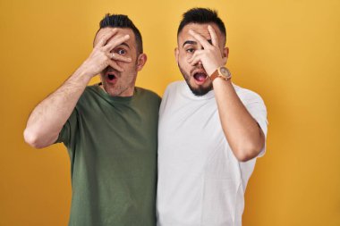 Homosexual couple standing over yellow background peeking in shock covering face and eyes with hand, looking through fingers with embarrassed expression. 