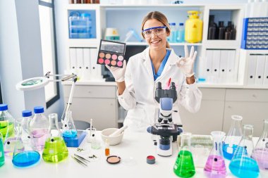 Bilim adamı laboratuvarında makyaj yapan genç sarışın kadın parmaklarıyla imza atıp gülümsüyor ve mükemmel bir sembol oluşturuyor. 