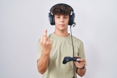 Hispanic teenager playing video game holding controller showing middle finger, impolite and rude fuck off expression 