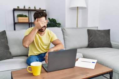 Young man with beard using laptop at home smiling and laughing with hand on face covering eyes for surprise. blind concept. 