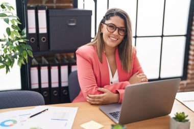 Young hispanic woman business worker using laptop sitting with arms crossed gesture at office