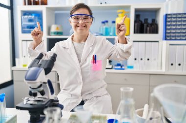 Down sendromlu İspanyol kız bilim adamı laboratuvarında çalışıyor. Şaşırmış, şaşırmış ve parmaklarıyla işaret ediyor ve kollarını kaldırıyor.. 