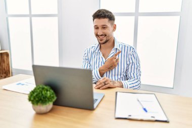 Ofiste çalışan sakallı yakışıklı genç adam bilgisayarda neşeli bir dizüstü bilgisayar kullanıyor. Yüzünde mutlu ve doğal bir ifadeyle, elleri ve parmaklarıyla yüzünü gösteren bir gülümseme var. 