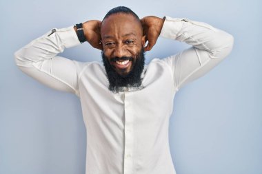 Afro-Amerikan adam mavi arka planda duruyor rahatlıyor ve esniyor, kolları ve elleri başının arkasında mutlu bir şekilde gülümsüyor. 
