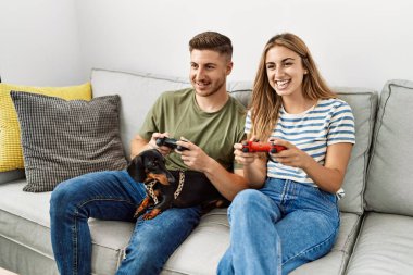 Genç İspanyol çift gülümseyerek evde köpekle kanepede oturup video oyunu oynuyor..