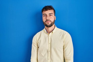 Handsome hispanic man standing over blue background relaxed with serious expression on face. simple and natural looking at the camera. 