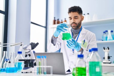 Laboratuvarda dizüstü bilgisayarda test tüpü tutan bilim adamı üniforması giyen İspanyol genç adam.