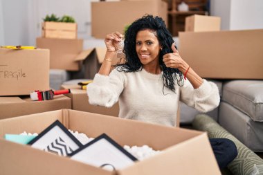 Orta yaşlı İspanyol bir kadın elinde mutlu ve mutlu bir şekilde gülümseyen, başparmağıyla mükemmel ve onay işareti yapan bir ev anahtarı tutuyor. 