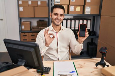 Sakallı genç İspanyol bir adam, küçük bir iş yerinde çalışıyor. Akıllı telefon ekranını gösteriyor. Parmaklarıyla imza atıyor, gülümsüyor. 