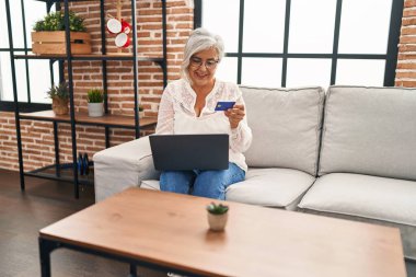 Evdeki koltukta oturan orta yaşlı bir kadın dizüstü bilgisayar ve kredi kartı kullanıyor.