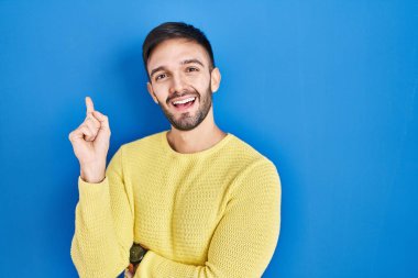 Mavi arka planda duran İspanyol bir adam yüzünde kocaman bir gülümsemeyle, kameraya el ile işaret ederek bakıyor.. 