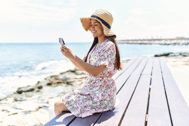 Yaz tatili giymiş, akıllı telefon kullanan genç Latin kız sahildeki bankta oturuyor..