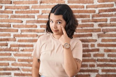 Young hispanic woman standing over bricks wall pointing to the eye watching you gesture, suspicious expression 