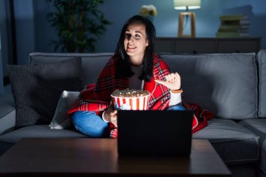 Hispanic woman eating popcorn watching a movie on the sofa smiling with happy face looking and pointing to the side with thumb up. 