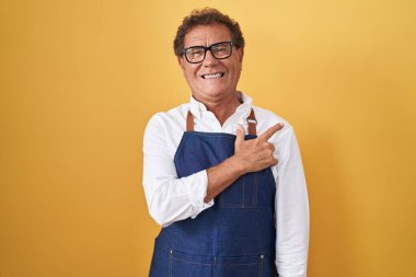 Middle age hispanic man wearing professional cook apron cheerful with a smile of face pointing with hand and finger up to the side with happy and natural expression on face 