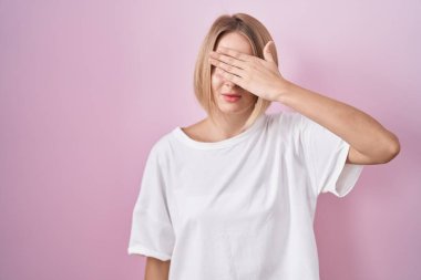 Young caucasian woman standing over pink background covering eyes with hand, looking serious and sad. sightless, hiding and rejection concept 