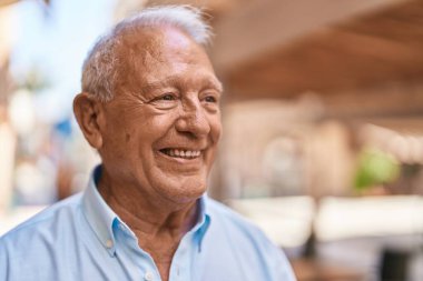 Yaşlı, kır saçlı, gülümseyen, kendine güvenen bir adam sokakta duruyor.