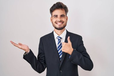 Dövmeli, takım elbiseli, kravatlı, avuçlarını gösteren ve baş parmağıyla hareket eden mutlu ve neşeli gülümseyen İspanyol bir genç. 