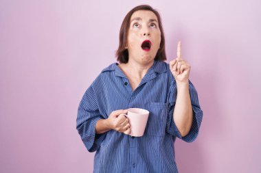 Orta yaşlı Latin bir kadın kahve içerken şaşkın şaşkın yukarı bakıyor ve parmaklarıyla işaret ediyor ve kollarını kaldırıyor.. 