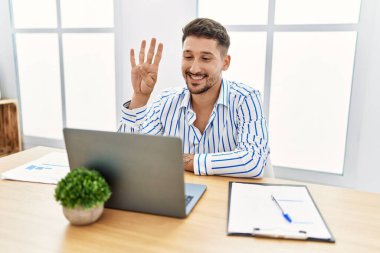 Ofiste çalışan sakallı yakışıklı genç adam bilgisayarını kullanarak dört numarayı gösteriyor ve gülümsüyor, kendinden emin ve mutlu.. 