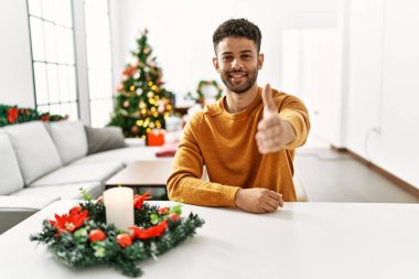 Arap genç adam Noel ağacının yanında masada oturuyor. Dostça gülümsüyor. Karşılama ve hoş geldin demek için tokalaşıyor. başarılı iş. 