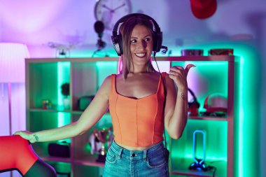Young woman playing video games wearing headphones pointing thumb up to the side smiling happy with open mouth 