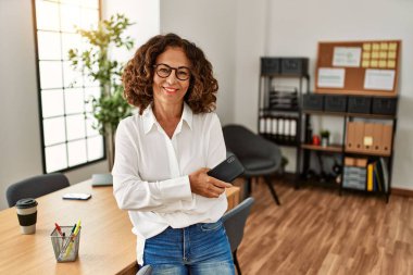 Ofiste akıllı telefon tutan orta yaşlı İspanyol kadın gülümsüyor.