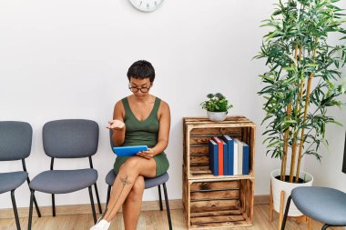 Young hispanic woman having video call sitting on chair at waiting room