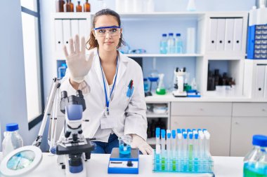 Young hispanic woman working at scientist laboratory doing stop sing with palm of the hand. warning expression with negative and serious gesture on the face. 