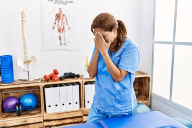 Middle age hispanic physiotherapist woman working at pain recovery clinic with sad expression covering face with hands while crying. depression concept. 
