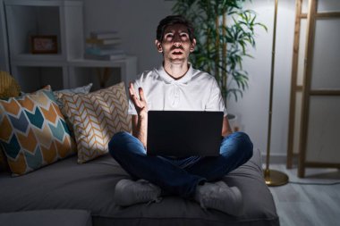 Young hispanic man using laptop at home at night crazy and mad shouting and yelling with aggressive expression and arms raised. frustration concept. 