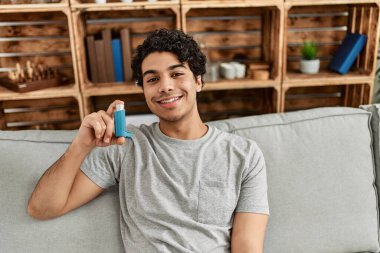 Evdeki kanepede oturan İspanyol asıllı genç adam astım spreyi kullanıyor..