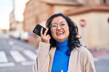 Young chinese woman smiling confident listening audio message by the smartphone at street