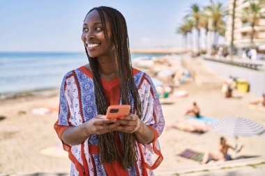 Genç Afrikalı Amerikalı kadın sahilde akıllı telefon kullanıyor..