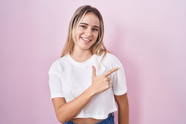 Young blonde woman standing over pink background cheerful with a smile on face pointing with hand and finger up to the side with happy and natural expression 