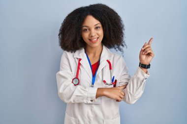 Genç Afro-Amerikan kadın doktor üniforması giyiyor ve steteskop takıyor. Yüzünde kocaman bir gülümseme var. Elleriyle kameraya bakıyor.. 