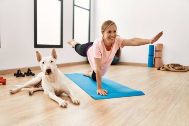 Genç beyaz kadın gülümsüyor spor merkezinde köpekle esniyor.