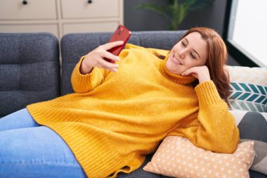 Young beautiful plus size woman using smartphone lying on sofa at home