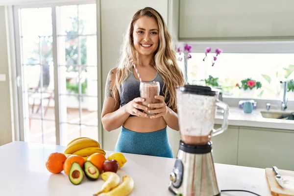 Young woman smiling confident holding glass of smoothie at kitchen