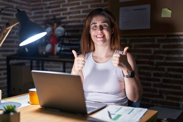 Brunette woman working at the office at night success sign doing positive gesture with hand, thumbs up smiling and happy. cheerful expression and winner gesture.