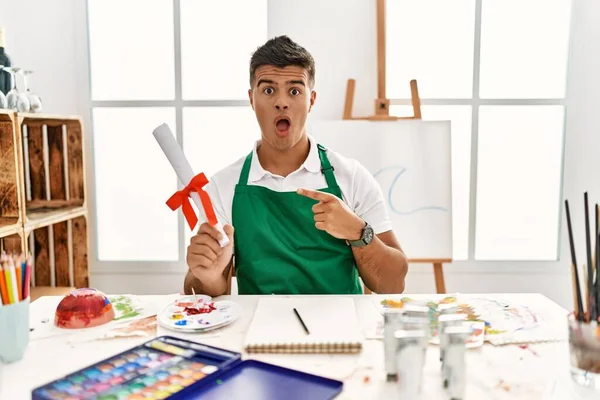 Joven Hombre Hispano Estudio Arte Sosteniendo Grado Sorprendido Señalando Con — Foto de Stock