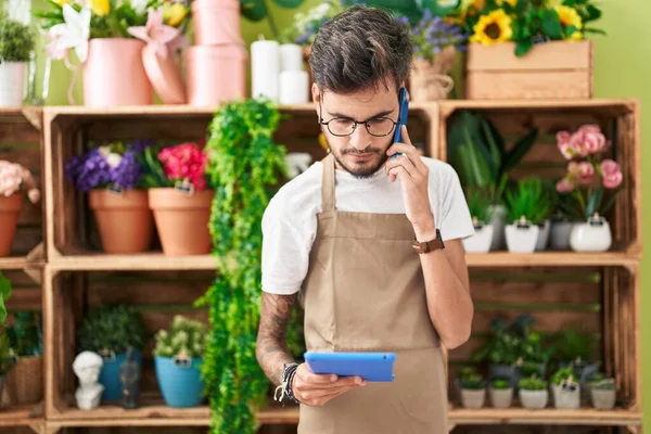 Young hispanic man florist talking on smartphone using touchpad at flower shop