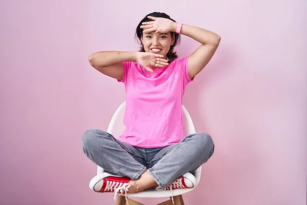 Hispanic young woman sitting on chair over pink background smiling cheerful playing peek a boo with hands showing face. surprised and exited