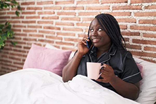 African american woman talking on smartphone drinking coffee at bedroom