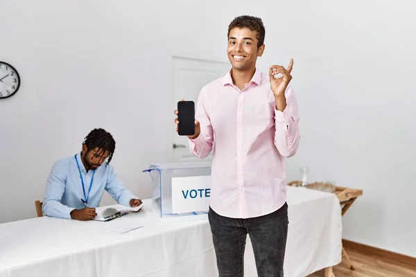 Young hispanic men at political campaign election holding smartphone smiling happy pointing with hand and finger to the side