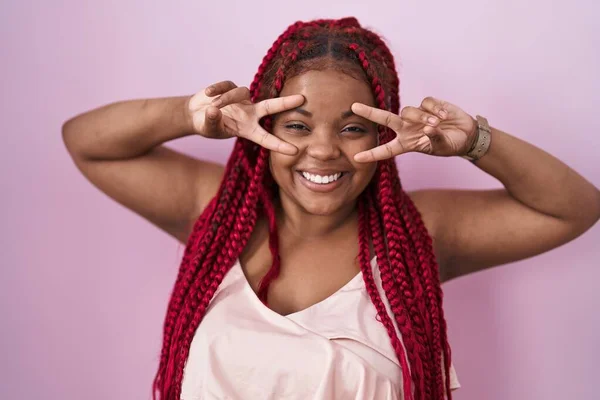 髪の毛を編んだアフリカ系アメリカ人女性がピンクの背景の上に立って顔の上に指で平和のシンボルを行います 勝利を示す明るい笑顔 — ストック写真