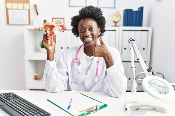 African doctor woman holding anatomical model of female genital organ at medical clinic smiling happy and positive, thumb up doing excellent and approval sign