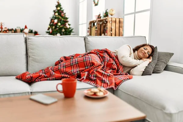 Middle age hispanic woman sleeping lying on the sofa by christmas tree at home.