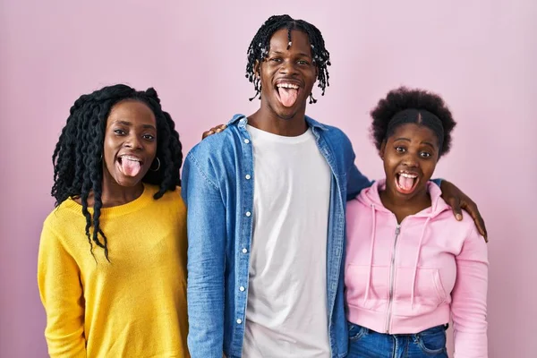 Group of three young black people standing together over pink background sticking tongue out happy with funny expression. emotion concept.