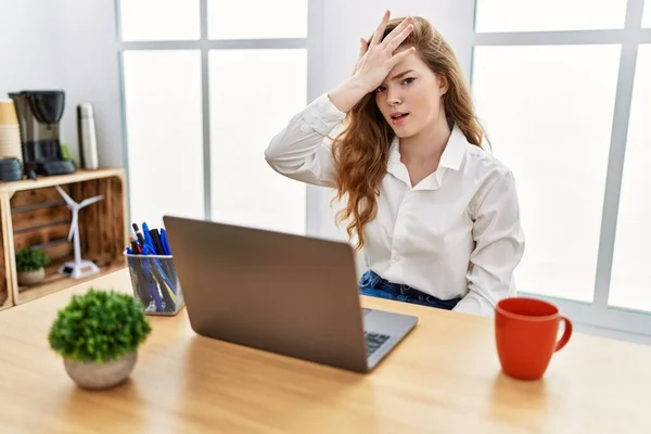 Young caucasian woman working at the office using computer laptop surprised with hand on head for mistake, remember error. forgot, bad memory concept.